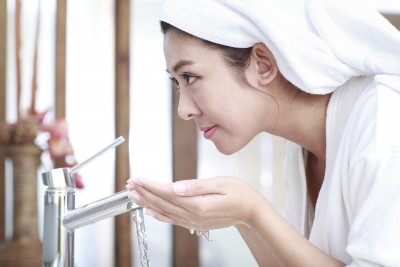 Woman wearing a towel on hear head, leaning over a sink and cupping water in her hands.