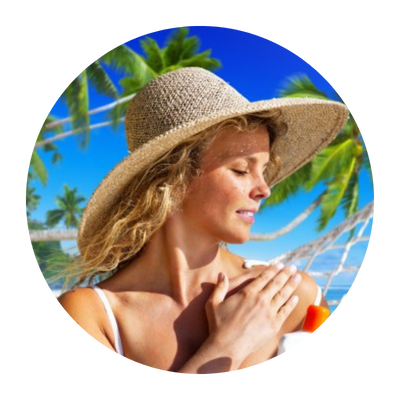 Woman in a wide brimmed hat applying sunscreen on a beach in front of green palm trees.