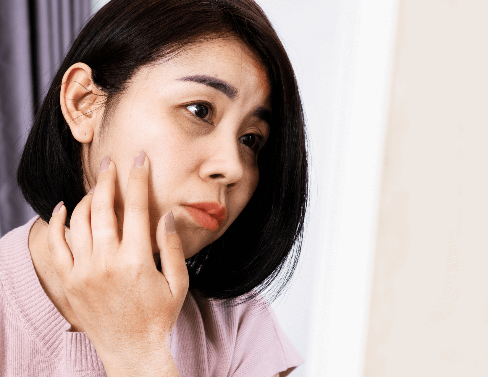 Woman looking in the mirror with a sad expression, while holding her hand to her face.