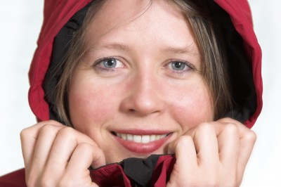 Woman's face up close, wearing a red rain coat with the hood up, smiling and holding the collar of her jacket with both hands. 