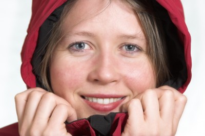 Visage de femme de près, portant un imperméable rouge avec la capuche relevée, souriant et tenant le col de sa veste à deux mains.