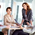 Des femmes dans un bureau lumineux, assises autour d’un bureau, souriant et buvant dans des tasses tout en travaillant. Une femme est debout.