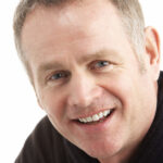 Man's face up close, smiling at the camera, against a white background.