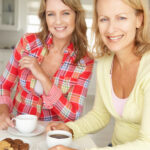 Two women sitting at a table, drinking out of mugs, smiling.