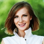 Woman with short hair blowing back in the wind, standing in front of a green background, smiling.